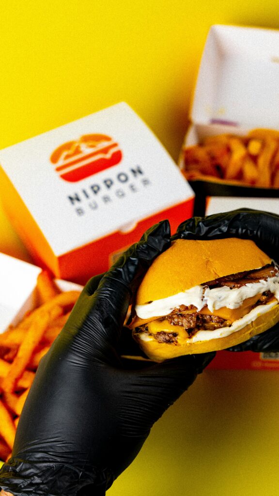 Close-up of a cheeseburger held in black gloves with fries on a bright background.
