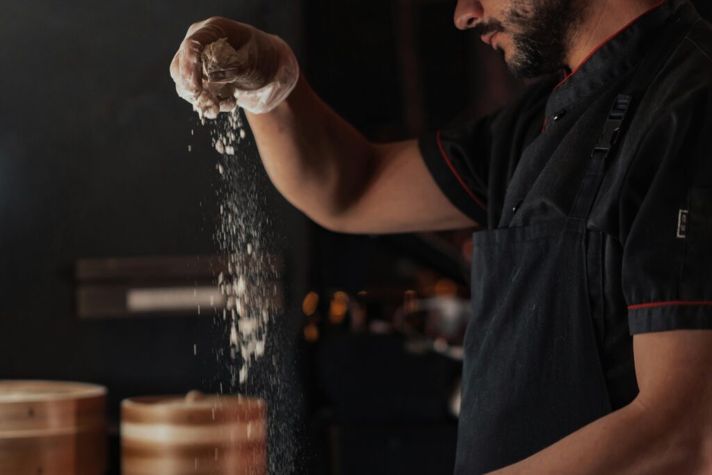 A chef wearing a black apron sprinkles flour onto a surface in a dimly lit kitchen, showcasing food preparation.
